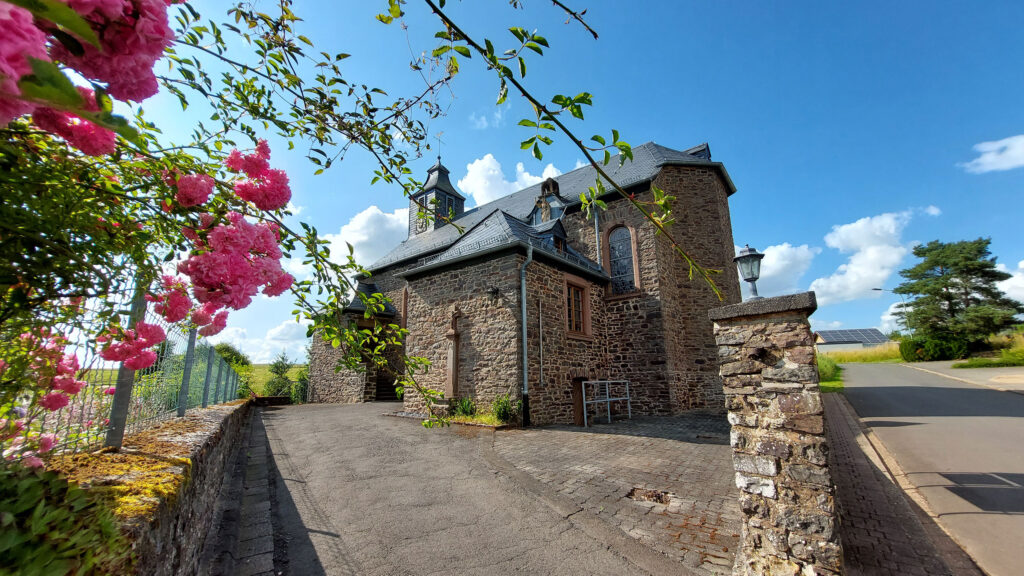 Historische Kapelle in Wawern bei Sonnenschein mit Natursteinmauerwerk und blühenden Rosen im Vordergrund – idyllisches Kulturerbe im Eifelkreis Bitburg-Prüm.