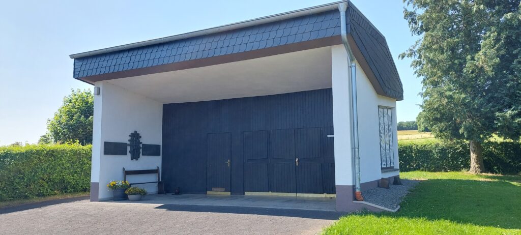Friedhofskapelle in Wawern bei Sonnenschein – moderner Bau mit dunkler Holztür, Gedenktafeln, Blumenbank und gepflegtem Außengelände im Grünen